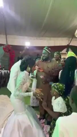 The video shows a newlywed couple in a reception area surrounded by guests, all dancing. The bride is wearing a white lace wedding dress, and the groom is in a lighter brown tunic and cap. Some guests are dressed in traditional African clothing with bright colors and patterns. The room is decorated with green and red draping, and there are some rose decorations and a table with bottles visible. The lighting shifts between white and green. Guests toss what appears to be money towards the couple as they dance.