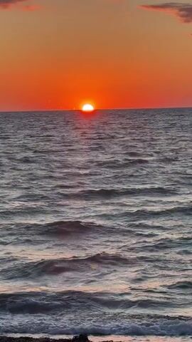 The video shows a sunset over the ocean. The sun is a bright orange disk partially submerged below the horizon. The sky is a gradient of orange and pink. The ocean is a deep blue-gray with gentle waves. The foreground shows a rocky shoreline. The video is about 5 seconds long and the camera is stationary. There are no people or objects visible in the video other than the natural elements of the sunset, ocean, and shoreline.