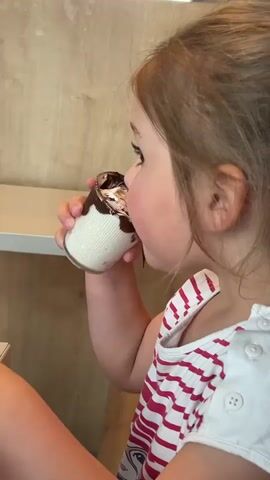 A young girl with light brown hair and wearing a white and red striped tank top is seen in profile, drinking from a small glass filled with white cream and chocolate sauce. She tips the glass up to her mouth and drinks, then sets the glass down. The background consists of a light wood-paneled wall and a light wood table.  A white countertop is visible on the left. The camera angle is slightly above her, capturing her face and upper body.