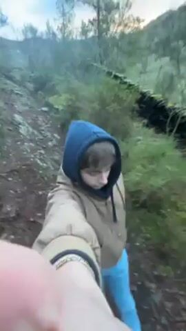 A young person, appearing male, is walking on a narrow, dirt path. They are wearing a tan jacket with a blue hood, light blue pants, and a visible gold bracelet. The camera is held in front of them in selfie style, with a portion of the hand visible in the foreground. The path is lined with greenery and an old stone wall on the right. Trees are visible in the background. The sky is overcast. The person makes a blowing sound, perhaps imitating the wind.