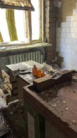 The video shows the interior of an abandoned room in disrepair. A window with a view of trees is on the left, with a radiator beneath. A table stands by the window, cluttered with debris, a couple of brown bottles, and a test tube rack. A row of drawers is partially open. To the right of the table is a large, deteriorating, red-topped counter with a green-and-white tiled edge. The white-tiled wall behind the counter shows stains of red and green. The room is littered with debris, paint chips, and broken pieces. The overall impression is one of decay and abandonment.