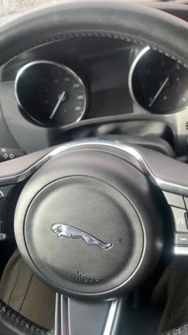 The video shows the interior of a car on a rainy day. The steering wheel with a Jaguar logo is visible, as well as the dashboard, window controls, and a side mirror. Raindrops cover the windows, obscuring the view of buildings outside. The video ends with a view of the raindrops on the sunroof, with the sun shining through. A sound is heard as the video comes to a close.