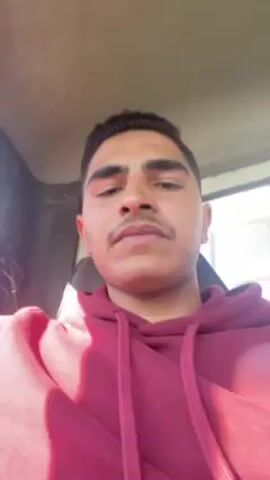 A young man with a mustache wearing a burgundy hooded sweatshirt is in what appears to be a car. He is looking at the camera and smiling and talking. He appears to be inside the car. The lighting is bright and sunny.