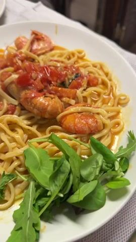 The video shows a close-up view of a plate of spaghetti with shrimp and tomato sauce. The spaghetti is arranged in a nest on a white plate, topped with several large shrimp and a chunky tomato sauce. A small garnish of green leaves, possibly arugula, is placed on the side of the plate. In the background, another plate of the same dish is partially visible. The lighting is warm and soft, suggesting an indoor setting, possibly a restaurant. The camera movement is subtle, with slight pans and zooms that focus on the texture and colors of the food.