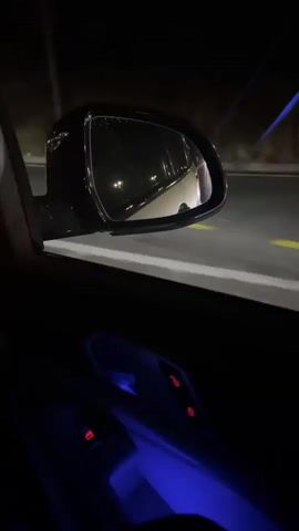A first-person perspective video shows the interior of a car at night. The camera is focused on the side mirror and the door panel. In the side mirror, lights from street lamps and passing cars are reflected. The door panel has ambient blue lighting and red buttons for locking and unlocking doors. The video appears to capture a moment of driving through a well-lit urban or suburban area at night.