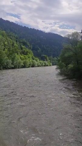 A wide, fast-flowing river dominates the foreground of the image. The water appears a murky brown and is agitated, with ripples and small waves visible. On the left side of the river, a steep, heavily forested mountainside rises dramatically. The trees are a vibrant green, and the foliage is dense. The mountain slopes upward towards a cloudy sky. On the right side of the river, the bank is lined with trees and vegetation, which appear slightly less dense than on the left. The sky is overcast with fluffy, greyish-white clouds, suggesting a possibly humid or rainy day. The overall impression is one of a wild, natural landscape.