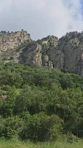 The video shows a pan of a mountainous landscape. The foreground is filled with lush green trees and foliage, with some distant vegetation. In the background, a large, rugged mountain face dominates the scene. The mountain is made of rock and appears steep and uneven, with some vegetation clinging to its slopes. The sky above is a mix of white clouds and blue sky. The camera pans slowly from left to right, revealing more of the mountain range and the surrounding greenery. There are no people or man-made objects visible in the video. The overall impression is one of a natural, wild, and untouched environment.