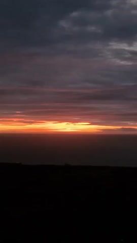 The video shows a dark silhouette of a landscape with sheep grazing on a field during a sunset. The sky is filled with dark clouds that are tinged with orange and pink from the setting sun. The sun appears as a bright, thin line just above the horizon. There are a few small structures visible in the distance. The overall mood is peaceful and serene.