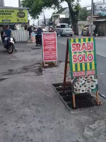 The video shows a street scene with several signs. In the foreground, there is a wooden A-frame sign advertising "SRABI SOLO" with an image of food below. To the left of this sign, there is another red sign with white text listing food items. Both signs are placed on a sidewalk next to a street. People on motorcycles are visible in the background, along with other signs and buildings. The ground is paved and appears wet in some areas. Traffic is moving on the street.