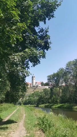 The video shows a view of a town with a prominent church or tower on a hill. The scene is a sunny day with a clear blue sky. In the foreground, there is a dirt path running alongside a body of water, possibly a river. Lush green trees and vegetation are abundant on both sides of the path and water. The path appears to be a walking or cycling trail. There are a few small figures walking on the path in the distance. The town on the hill is white with a distinctive tower. The overall scene depicts a peaceful, scenic outdoor environment.