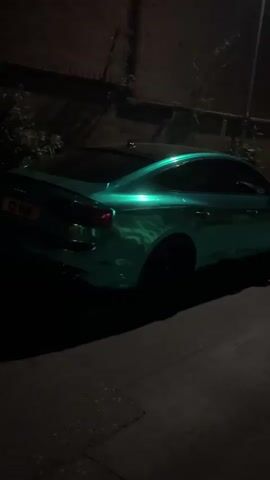 The image displays a sleek, modern car, likely an Audi A5 or S5 Sportback, parked on a street at night. The car is the primary subject and stands out due to its highly reflective, iridescent green or teal finish, which appears to be a custom wrap or paint job. Its surface gleams, catching and reflecting the available light. The vehicle has dark tinted windows and what appear to be black alloy wheels, partially obscured by shadows.

The scene is dimly lit by a single street lamp, visible on the right side of the frame, casting a warm glow and creating prominent reflections on the car's body. The ground beneath the car is dark asphalt. The background is largely dark and indistinct, showing what appears to be a wall or fence with some sparse, shadowy foliage. To the far right, behind the main car, a small portion of another dark vehicle is visible, including a partial UK license plate that begins with "DNZ" followed by "0_P". The overall impression is a quiet, nighttime street setting, with the unique car being the focal point. No people or specific activities are visible.