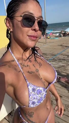 A woman wearing a purple and white floral bikini and sunglasses poses for a selfie on a wooden boardwalk at a beach. She has wet dark hair and multiple tattoos on her chest and abdomen. The ocean and other beachgoers are visible in the background.