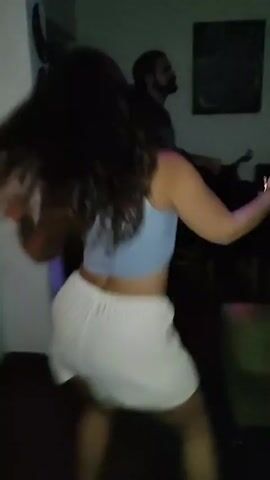 The video depicts two women dancing indoors, seemingly at a party or social gathering. The first woman, with curly brown hair, is wearing a light blue crop top and white shorts. She holds a phone, presumably recording the scene. A man with a beard stands blurry in the background. The second woman, who appears later in the video, is wearing a white crop top and denim shorts. She is dancing with a more free-flowing energy. The setting appears to be a simple living room or similar space, with neutral walls, a dining table in the background, and some decorative wall hangings. The room is dimly lit, with what appears to be a light effect, possibly from a DJ setup.