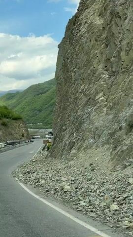 The video shows a roadside view, likely filmed from a moving car. A large rock face towers over the road, with loose rocks scattered on the asphalt. A road sign indicating "ANANURI 1000M" in both English and Georgian is visible. In the background, green hills and mountains complete the landscape.  A glimpse of Ananuri fortress can be seen behind the sign. The sky is partly cloudy with blue patches.