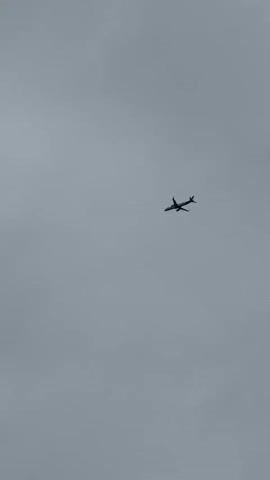 A video shows an airplane flying in a gray, overcast sky. The plane is small and distant, appearing as a dark silhouette against the uniform gray background. There are no other visible objects or people in the frame.