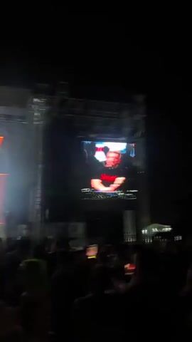 A nighttime outdoor concert is underway. A large screen displays a man at a DJ booth wearing a black t-shirt with red lettering. The screen is illuminated by stage lighting, which also casts beams through smoke or haze into the dark sky. A crowd of people stands in front of the stage, silhouetted against the lights. Some individuals are holding up their phones. The overall atmosphere is lively and energetic.