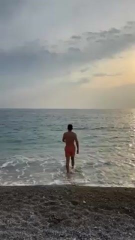 A man in red swim trunks walks into the ocean. The beach is composed of small gray pebbles. The ocean is calm with small waves. The sky is cloudy with a patch of light from the setting sun. The man is facing away from the camera.