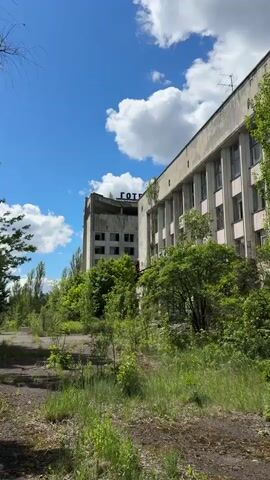 The video shows an overgrown, abandoned hotel with a sign on top that says "Hotel" in Russian, under a blue sky with fluffy white clouds. Greenery fills the foreground, with grass, weeds, and young trees obscuring the walkway leading to the hotel. The building is several stories tall and shows signs of disrepair, with broken windows and a weathered facade. Someone is walking along a narrow exterior walkway on the building's facade. The overall impression is one of desolation and nature reclaiming a man-made structure.