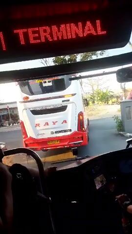 The video shows the interior of a bus, looking forward through the windshield. Above the windshield, a red LED display shows "1 TERMINAL". In front of the bus, another bus is visible, with its rear end facing the camera. This bus is white with red accents and has "RAYA" written on its back. It appears to be either parked or moving slowly. The environment appears to be outdoors, possibly a bus terminal or a road, with vegetation and structures visible in the background. The lighting suggests it might be daytime. A person's hand is visible on the steering wheel of the bus from which the video is being recorded.