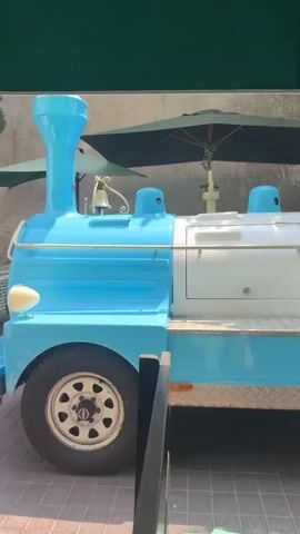 The video shows the side view of a blue and white train-like vehicle. The front of the vehicle appears to be a driver's cabin, with a driver and a passenger visible through the windows. The train has several passenger cars behind the driver's cabin, and there are multiple passengers in these cars. The vehicle is parked outdoors, with trees and umbrellas visible in the background. The ground is paved with tiles. The overall impression is of a tourist train.