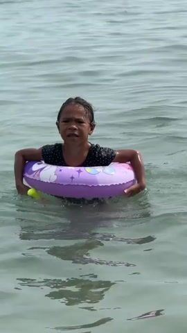 A short video shows a young Filipino girl swimming in the ocean, holding a purple and white unicorn-themed float. She has dark, wet hair and is wearing a black polka-dot swimsuit. She is trying to swim to the left, but the waves keep pushing her. Her facial expression alternates between determination and struggle. The weather seems to be sunny and the water is clear with visible reflections of the sky and the girl. The video seems to have been filmed in the Mampising area of the Philippines.