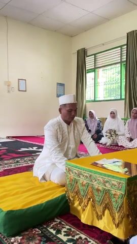 A man in a white ceremonial outfit and cap is kneeling on a yellow cushion. He is seated on a patterned carpet, facing a small, ornate table draped in yellow fabric with green and gold trim. In the background, three women in hijabs are seated. The woman in the center is holding a bouquet of white flowers. The room has light-colored walls and green curtains covering a window with green blinds. A framed picture hangs on the wall to the left. The lighting appears to be natural, possibly from the window. The scene suggests a formal or ceremonial gathering, likely a wedding or religious ceremony.