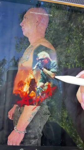 A person's torso is superimposed with a graphic image, possibly a tattoo design, featuring a man singing into a microphone. Flames and silhouettes of musicians are also visible in the background of the graphic. A hand with manicured nails holds a stylus, interacting with the image on a tablet screen. The background of the video appears to be outdoors, with greenery visible.