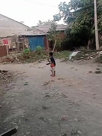 A young boy in a black t-shirt and tan shorts stands in a dirt area, holding a stick. He swings the stick as if playing a game. Behind him, there are some dilapidated structures with corrugated metal roofs and a blue tarp. To the right, a motorcycle is parked. Trees and foliage are visible in the background. The ground is uneven and dusty.