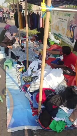 A street vendor stall displays used clothing for sale. The stall is set up outdoors, likely by the side of a road, indicated by the presence of a paved surface and a few vehicles and pedestrians in the background. The clothing is spread out on a large tarp, with some items hanging on a rack behind the tarp. Bamboo poles support a dark-colored tarp overhead, providing shade. The items for sale appear to be a mix of adult and children's clothing, with a variety of colors and patterns. Three people are visible at the stall. One person, wearing a hijab and a dark jacket, is seated and appears to be sorting through clothes. Another person, also wearing a hijab and a dark top, is seated and looking at clothing items. A third person, wearing a red shirt, is seated on the ground and handling clothes. A sign is visible behind the stall, featuring text and an image of a woman. The ground is covered with a blue and white striped tarp, on which the clothing is piled. The overall scene depicts a typical street market environment for second-hand goods.