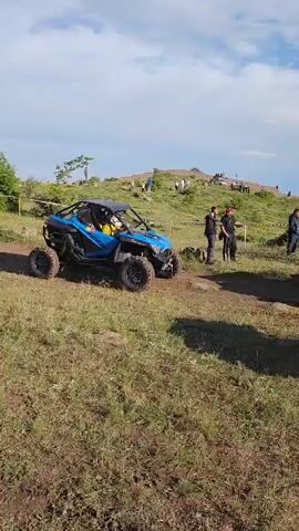 This video shows an off-road racing event taking place in Antarut, Armenia. The scene is set on a grassy, hilly terrain under a partly cloudy sky, suggesting it is daytime. Several individuals are present, some standing and observing, while others appear to be participants or event staff.

The main objects of interest are various off-road vehicles, specifically UTVs or side-by-sides. One prominent blue vehicle is seen in the foreground of the initial clip, while subsequent clips focus on a white and red vehicle with "RC" prominently displayed on its side. These vehicles are parked or moving on a dirt track.

The event appears to be at or near a starting line, indicated by a large archway with the word "START" clearly visible in blue text. Red flags and other banners are also visible, adding to the festive or competitive atmosphere of the event. The overall mood seems to be one of anticipation and excitement, typical of a race. The terrain is somewhat rough and natural, with dry grass and some loose dirt, suitable for off-road activities.