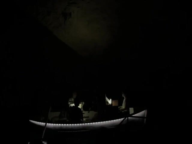 A small boat with a light along its edge floats in near-total darkness, with the shapes of four people visible. The space above them is a cavern ceiling with a light source shining from above. A single oar is visible at the front of the boat.