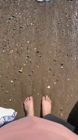 The video shows a pair of bare feet standing on a beach. Waves are crashing onto the shore, washing over the feet. The beach is covered in sand and small pebbles. The person is wearing a pink top and black pants. A small, patterned cloth is visible on the left side of the frame.