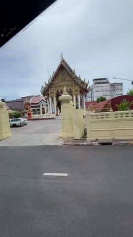 The video begins by showing a wide shot of a building, possibly a temple, with ornate golden detailing and tiered roofing. The building is situated behind a large gate with decorative pillars and a fence. A paved road is visible in the foreground. The camera then moves inside a vehicle, likely a bus. The interior of the bus is decorated with brown quilted leather on the ceiling and seats. Some seats are visible with purple patterned cushions. Passengers are seated in the bus. The camera pans across the interior, showing the dashboard, a backpack, and the windows. The video ends with a shot of the interior of the bus, showing more rows of seats and passengers.