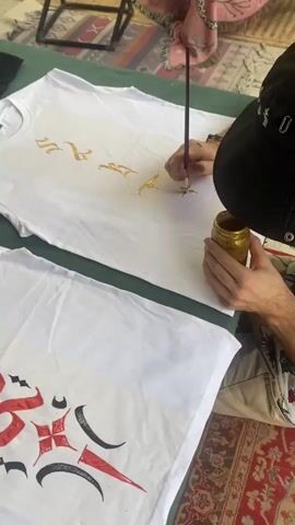 A person wearing a black bucket hat is seated, holding a small jar of gold paint and a paintbrush. They are painting gold details onto a white t-shirt with ornate gold lettering. Next to this t-shirt is another white t-shirt with a design in red, black, and gray. The scene is set on a patterned rug with a green and black underlay. A small black stool is visible in the background.