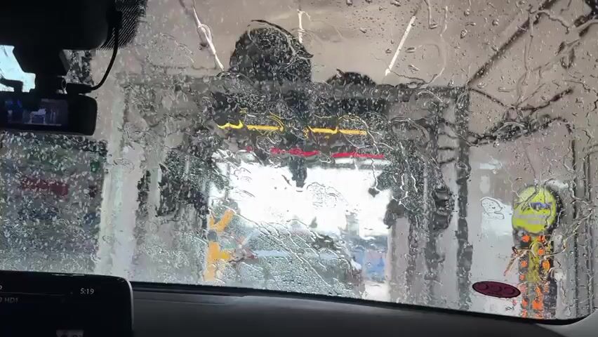 A car's windshield viewed from the interior, with water spraying and streaking down the glass, obscuring the view of a car wash tunnel. Through the water, parts of the car wash equipment, including brushes, lights, and a yellow sign with an orange spiral, are visible. A dashcam is mounted on the windshield near the rearview mirror. The car's dashboard with a display screen showing the time 5:19 is visible in the lower part of the frame.