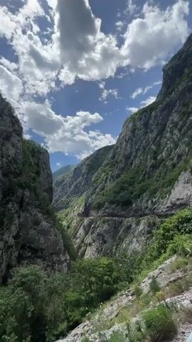 Here's a description of the video:

The video shows a scenic view of a mountainous canyon under a partly cloudy sky.  The steep, rocky cliffs are covered with patches of green vegetation. A narrow road winds through the canyon. The camera then tilts upwards to highlight the towering rock formations. Next, the camera shows a man sitting on a railing overlooking the canyon.  Cars are parked near a tunnel entrance.