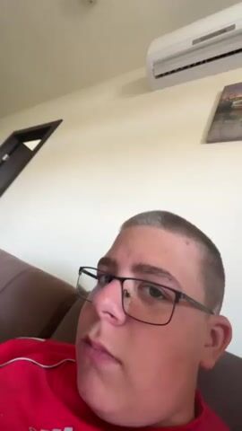 A young male with short hair and glasses is shown in a close-up video. He is wearing a red shirt. The background appears to be an indoor setting, possibly a living room, with a couch, a wall, and an air conditioning unit visible. The video is shaky and the camera angle is dynamic, suggesting the person is moving the camera around.