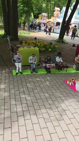 The camera pans across a street scene in what appears to be a park, where a group of animatronic musicians is set up along a pedestrian walkway. The walkway is paved with grey bricks. The musicians include a trumpet player, a saxophone player, a drummer, a pianist, a guitarist, and an accordion player. Two stuffed dogs are lying on a pink mat in front of the musicians, next to a white donation bucket. A man wearing a green long-sleeved shirt and a baseball cap places money into the donation bucket. In the background, there are colorful, whimsical buildings, possibly part of a market or festival, and people can be seen walking around. The area is surrounded by trees and greenery. The overall atmosphere is festive and lively.