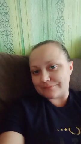 The video shows a close-up, selfie-style shot of a woman with her hair pulled back. She is wearing a dark t-shirt. She is sitting on a couch, and the background appears to be a wall with light green patterned wallpaper. The lighting is soft, and the woman is smiling slightly at the camera. The video is brief and shows no significant action or interaction.