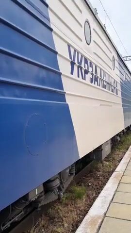 A passenger train is seen at a station platform. The train is white and blue. The text on the train reads "УЗ" in large blue letters with white outlining. The camera is moving alongside the train, showing the side of multiple carriages. There is a platform to the right of the train, made of square concrete tiles. In the distance, two people can be seen walking along the platform. The sky is overcast and there are hills in the background.