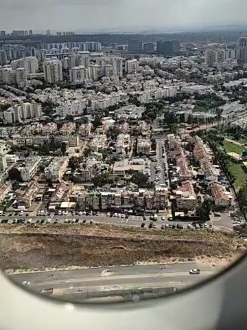 An aerial view captures a sprawling urban landscape from an airplane window. The city is densely populated with numerous apartment buildings and residential areas. In the foreground, a park with green grass, colorful structures, and recreational facilities is visible. Cars are parked along roads and in residential areas. The horizon is lined with taller buildings, suggesting a larger metropolitan area. The sky is partly cloudy. The video appears to be a continuous shot as the plane likely moves forward.