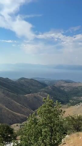 This is a vertical video capturing a scenic view from a high vantage point, likely a mountain or elevated viewpoint in the Kassopaia Municipal Unit, Greece. The scene transitions from a wide panorama of a serene, hazy body of water with distant, mountainous islands under a bright blue sky with scattered clouds, to a closer view of a rugged, stone-built structure.

Initially, the camera pans across a verdant landscape of rolling hills covered in trees and scrub, with winding paths visible. The shimmering water dominates the background, suggesting a large bay or sea. The overall impression is one of natural beauty and tranquility, with the clear, bright sky indicating it's daytime, likely late morning or early afternoon.

The scene then shifts focus to a stone building, possibly a church or monastery, constructed from rough, irregular white stones. A stone-paved courtyard and a low stone wall with a railing are prominent features. The building has arched doorways and small, arched windows with green shutters. The architecture suggests an older, possibly historic, structure. The sunlight casts shadows, further confirming the daytime setting. There is no visible text in the video. The mood conveyed is one of peaceful contemplation and admiration of both the natural landscape and the historical architecture.