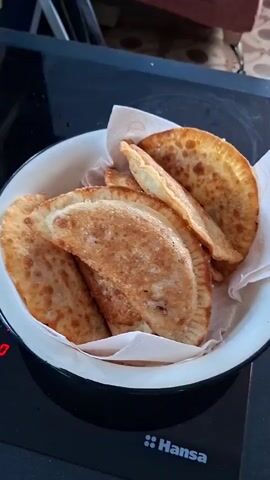 A close-up overhead shot shows a white enamel bowl filled with fried pastries. The pastries are golden brown, crescent-shaped, and appear to be stuffed. They are piled on top of each other, with a white paper napkin underneath the topmost ones. The bowl sits on a black stovetop, with the red digital display of a burner visible on the left side of the frame. The brand name "Hansa" is visible on the stovetop. The background is out of focus, but appears to be a kitchen environment with tiled flooring and furniture.