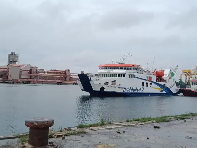 The video shows a ferry boat docked in a harbor. The ferry is predominantly white with a blue stripe along the hull, and orange accents on the upper deck. It is labeled "acehhebat 2."  A tugboat is positioned next to the ferry, possibly assisting with docking or maneuvering. The water in the harbor is relatively calm. In the background, there is industrial infrastructure, including large buildings and structures that suggest a port or factory. The sky is overcast, suggesting a cloudy day. A bollard is visible in the foreground, on what appears to be a concrete dock.