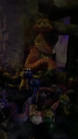 The video shows a dark, cavernous environment with rocky, uneven surfaces and artificial lighting. The focus is on a collection of colorful, puppet-like figures, likely part of a display or attraction. Prominently featured is a large, orange, serpent-like creature with a rounded head and prominent cheeks. Surrounding it are several smaller, diverse figures in blues, purples, and greens, some appearing to have animalistic features. The background includes dark, cave-like structures, with intermittent flashes of purple and blue light, suggesting an artificial lighting scheme designed to create atmosphere. The video appears to be handheld, with slight camera movement.
