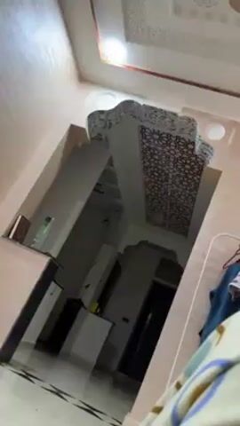 The video presents an interior shot of a room with noticeable architectural details. The walls appear to have a textured, possibly peach-colored finish. There is an arched opening with decorative trim at the top and edges, revealing a glimpse into a room with similar architectural features. The ceiling in both spaces is white with an ornamental pattern. At the side, a shelf-like structure holds a brown book with gold accents, positioned next to a clear glass bottle with a green label. The flooring has a shiny, tile-like appearance. The overall lighting appears subdued, casting shadows and contributing to the ambiance of the space.