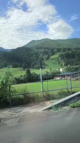 The video shows a view from a moving vehicle, looking out at a lush green landscape with rolling hills covered in trees. The sky is blue with scattered white clouds. In the foreground, there is a metal guardrail, and a fence is visible. The scene appears to be a rural or mountainous area, possibly in Europe given the architecture visible in the distance. There are no people or animals visible in the video. The video captures the serene beauty of nature.