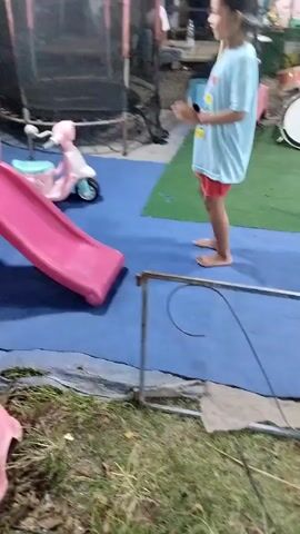 A young child in a light blue t-shirt and red shorts stands on a blue mat next to a pink plastic slide and a pink toy scooter. The child is facing right and appears to be mid-motion, possibly playing. Behind the slide and child, a trampoline is visible. In the foreground, a pink plastic stool is partially visible. The environment appears to be outdoors with grass and dirt.