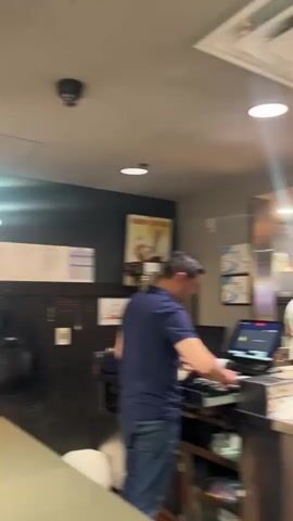 The video captures the interior of a casual restaurant or bar.

The first shot focuses on a service counter, where a man in a blue shirt is operating a point-of-sale system on the left. In the background, another man is visible working in a kitchen area. Above them, a large flat-screen TV is displaying a tennis match on what appears to be a clay court. The area is brightly lit with overhead lights and features stainless steel surfaces.

The camera then pans across the main dining area, revealing several patrons seated at tables and along a bar counter. The decor includes modern hanging light fixtures, patterned ceiling tiles, and abstract wall art. Red luggage can be seen near some of the seated individuals, suggesting a travel-related context, possibly an airport restaurant.

The final shot provides a close-up of a meal placed on the bar counter. The plate contains Mexican-style food, including a portion of orange rice, a small bowl of refried beans, and several folded quesadillas served with a side of sour cream. Next to the plate are a red can of Coca-Cola, a bottle of hot sauce, a salt shaker, and a mobile phone. A hand holding a white napkin is also visible in the frame, suggesting someone is about to eat or has just started their meal. The overall atmosphere appears relaxed and functional.