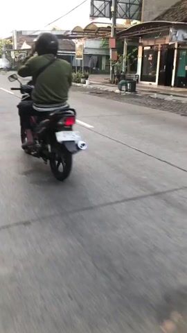 A first-person perspective video shows a motorcycle rider on a paved road with buildings lining the right side of the street. Another motorcycle rider is visible ahead in the same lane. The road has painted white lines and appears to be in a residential or commercial area. The sky is overcast.